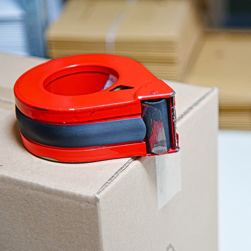 A red and black tape dispenser sitting on top of a brown cardboard box, with more stacked boxes blurred in the background.